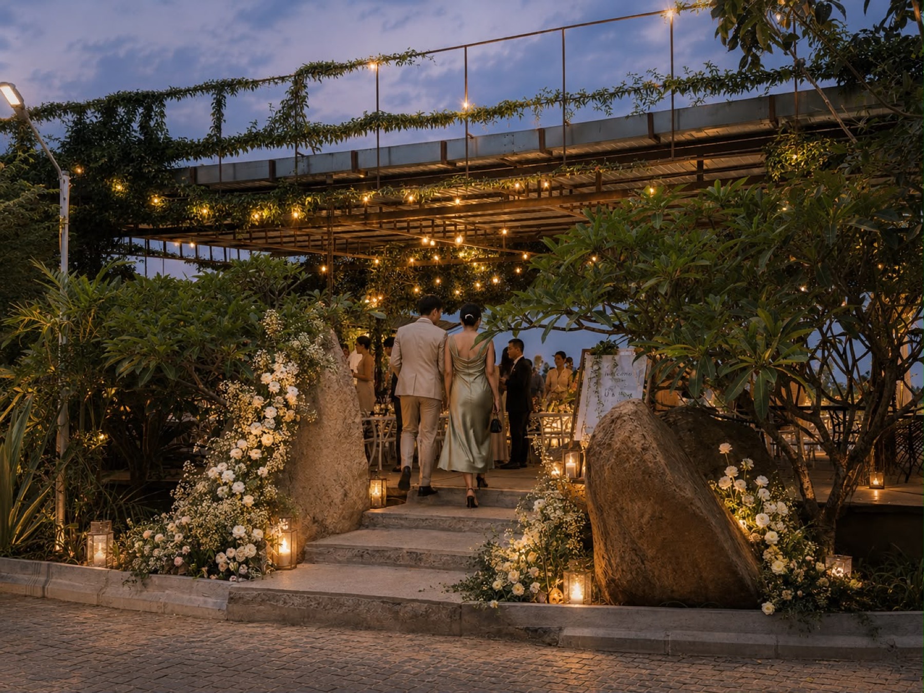 A couple walk up candlelit garden steps toward their ceremony at Orchid Tree, flanked by white flowers and greenery